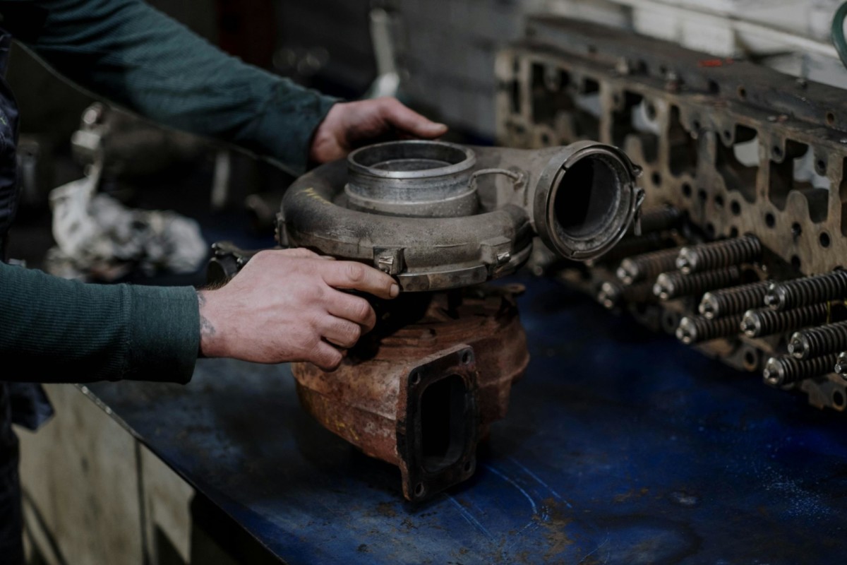 A person holding a mechanical part in a workshop, with a disassembled engine block and other parts on a table.