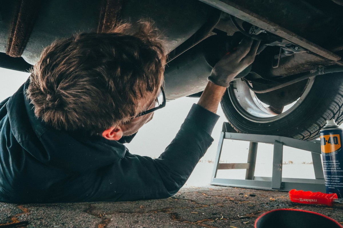 A man works on a car, lying on his back and reaching up into its undercarriage, with tools nearby on the ground.