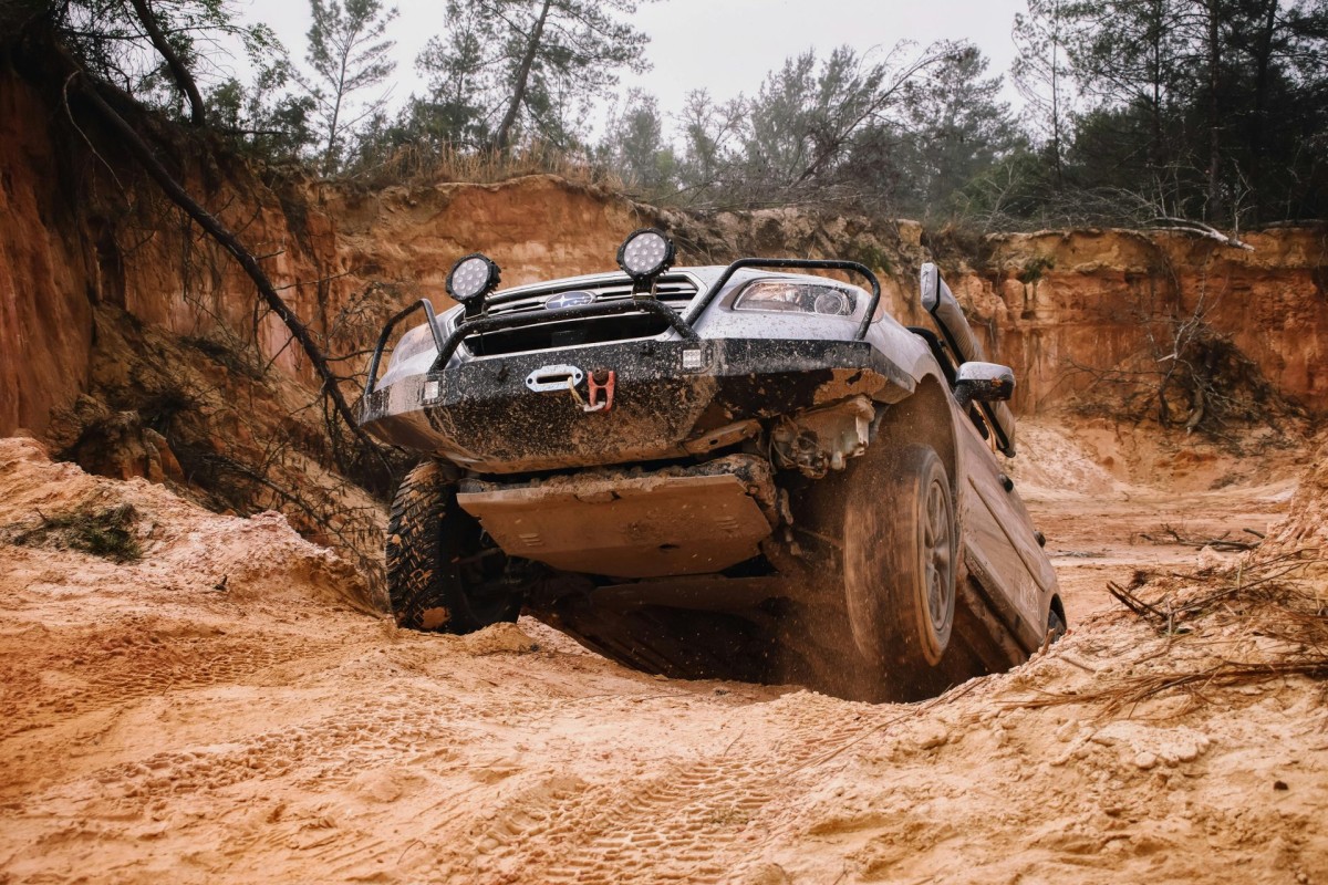 A black off-road vehicle, showcasing its car suspension, is tilted on uneven sandy terrain in a rugged outdoor area with trees and exposed soil.