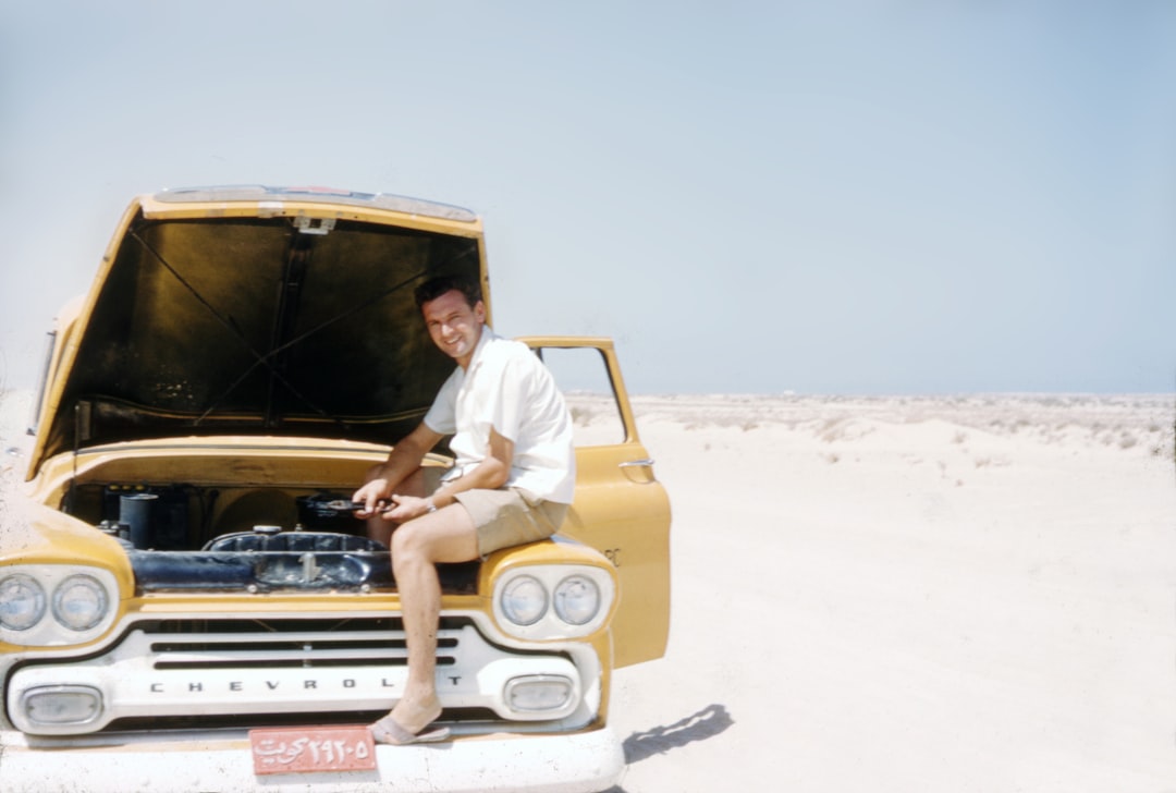 A man in shorts and a white shirt works under the hood of a yellow Chevrolet truck, performing a car battery replacement in a sandy, desert-like area under a clear sky.