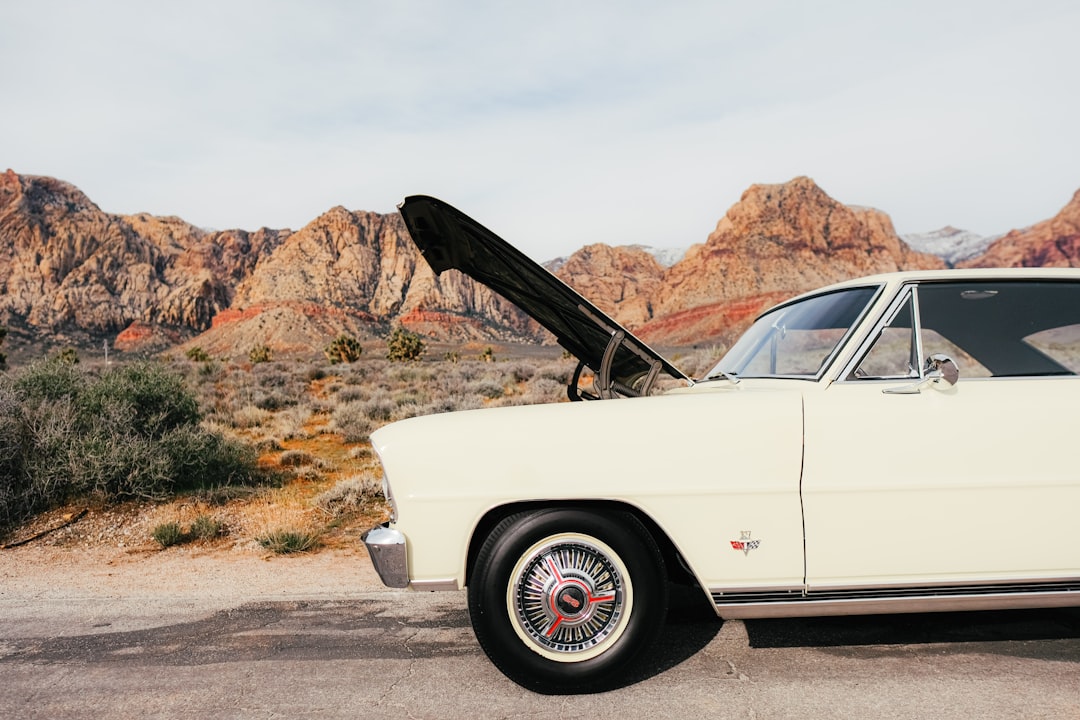 Home | Sartorial Auto Repairs A vintage cream-colored older car with its hood open is parked on a road in front of a desert landscape with rocky mountains, possibly awaiting a professional fuel system check.